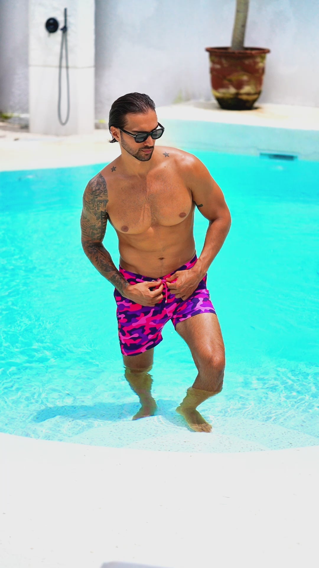 Man in pink camo swim shorts walking through a pool, wearing sunglasses.