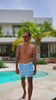 Man in blue swim trunks with standing poolside with palm trees in the background.