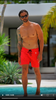 Man in bright red swim trunks standing by a poolside with palm trees.