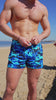 Man in blue camo swim trunks adjusting his waistband on the beach.