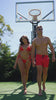 Couple in matching red swimwear walking on a basketball court with a hoop in the background.