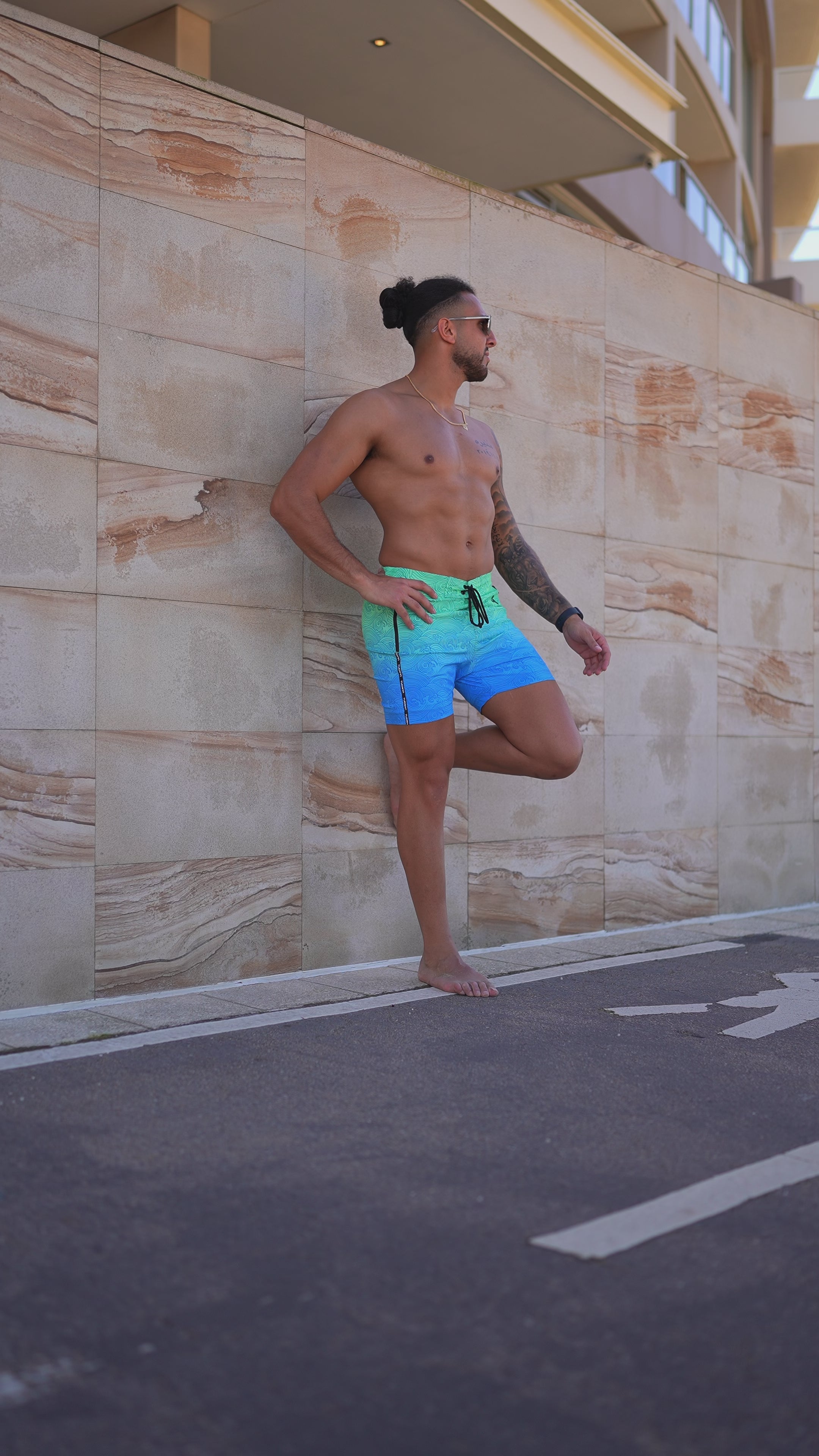 Man in turquoise-to-blue gradient swim trunks with faded waves design, leaning against a wall.