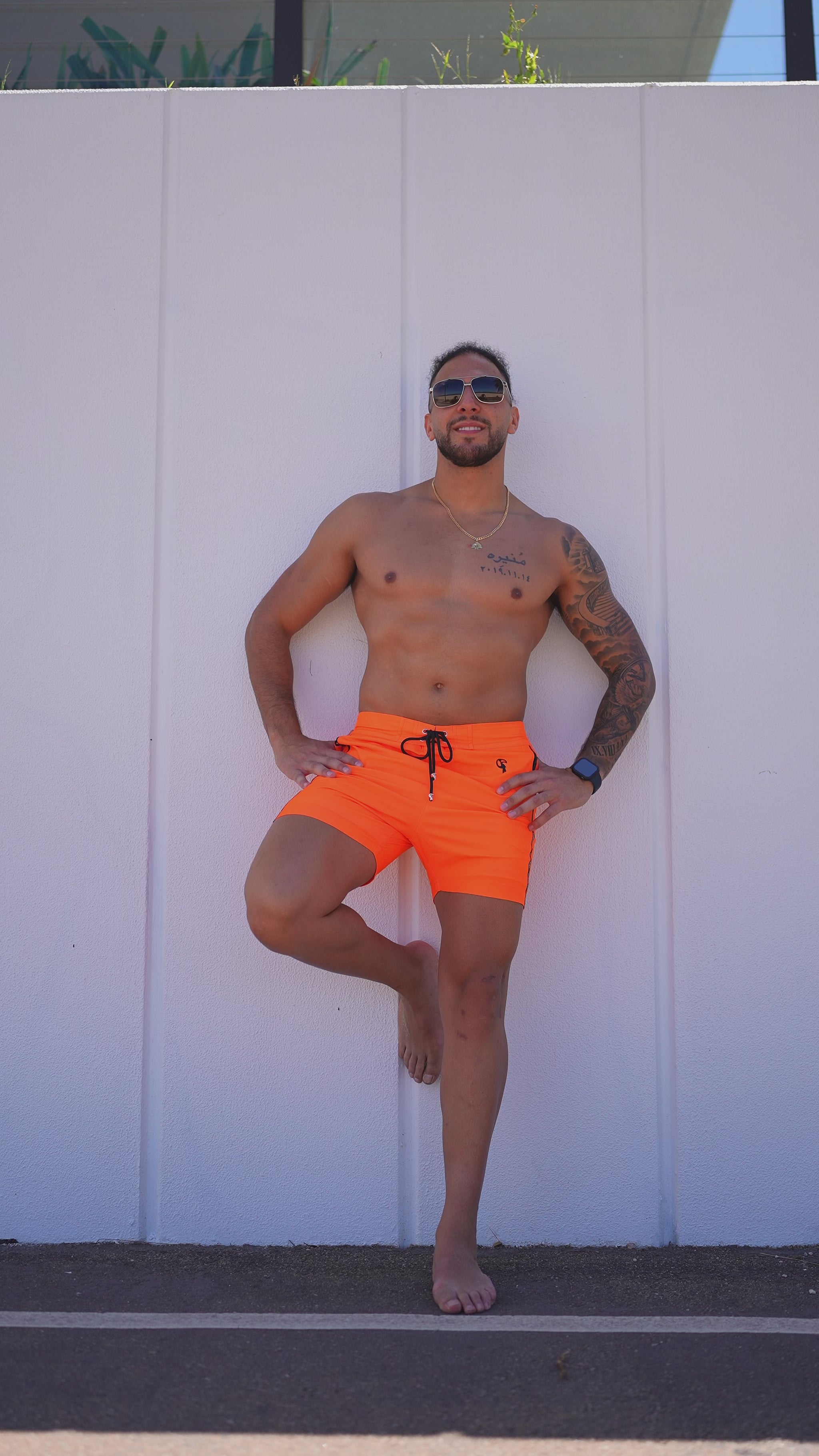 Man in bright orange swim trunks with a palm tree design posing against a white wall.