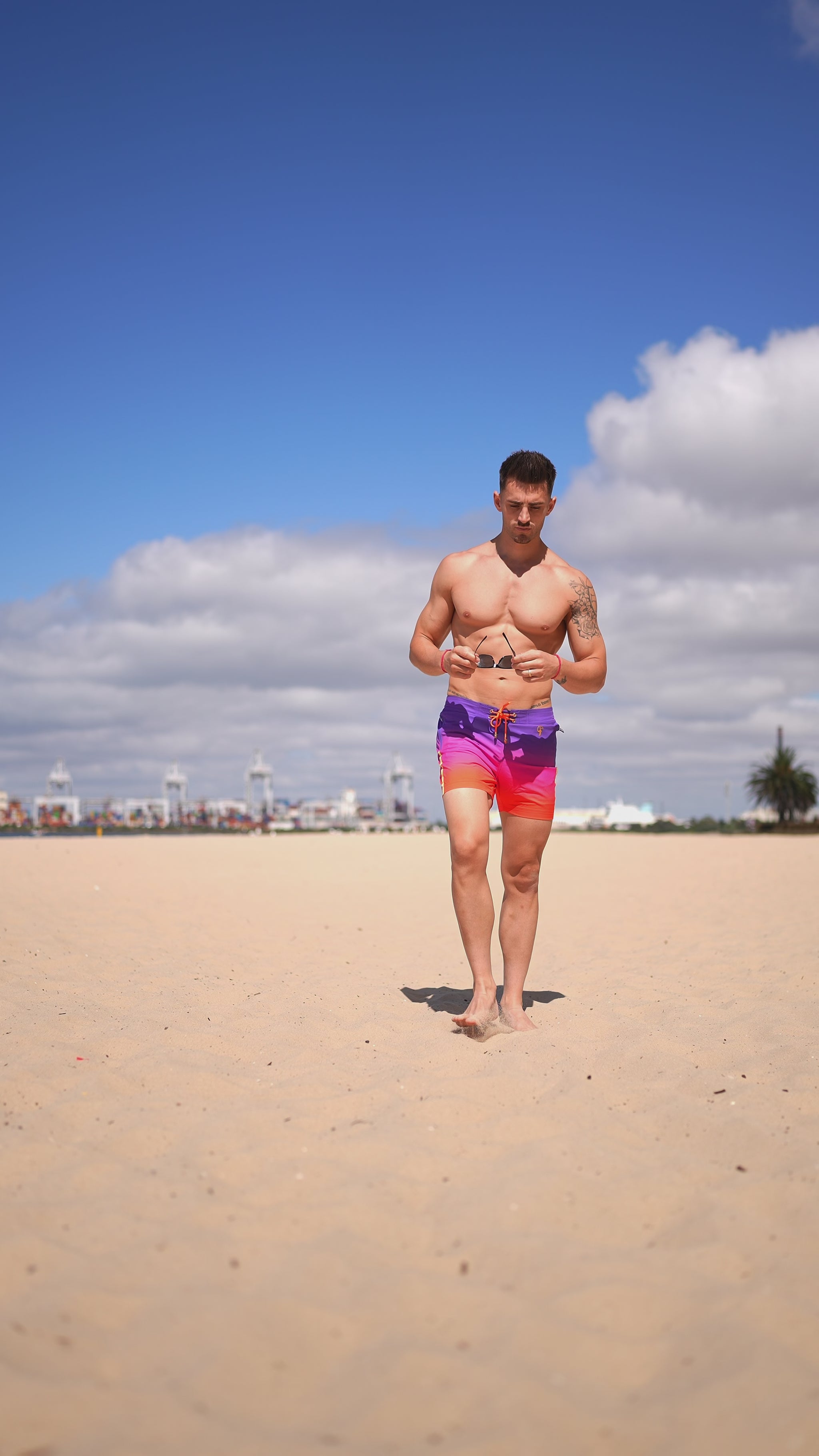 Man in faded crimson swim trunks walking on a sandy beach under a blue sky.