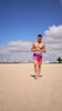 Man in faded crimson swim trunks walking on a sandy beach under a blue sky.