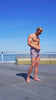 A man in cherry and cake print swim trunks poses by a coastal walkway under a clear blue sky.