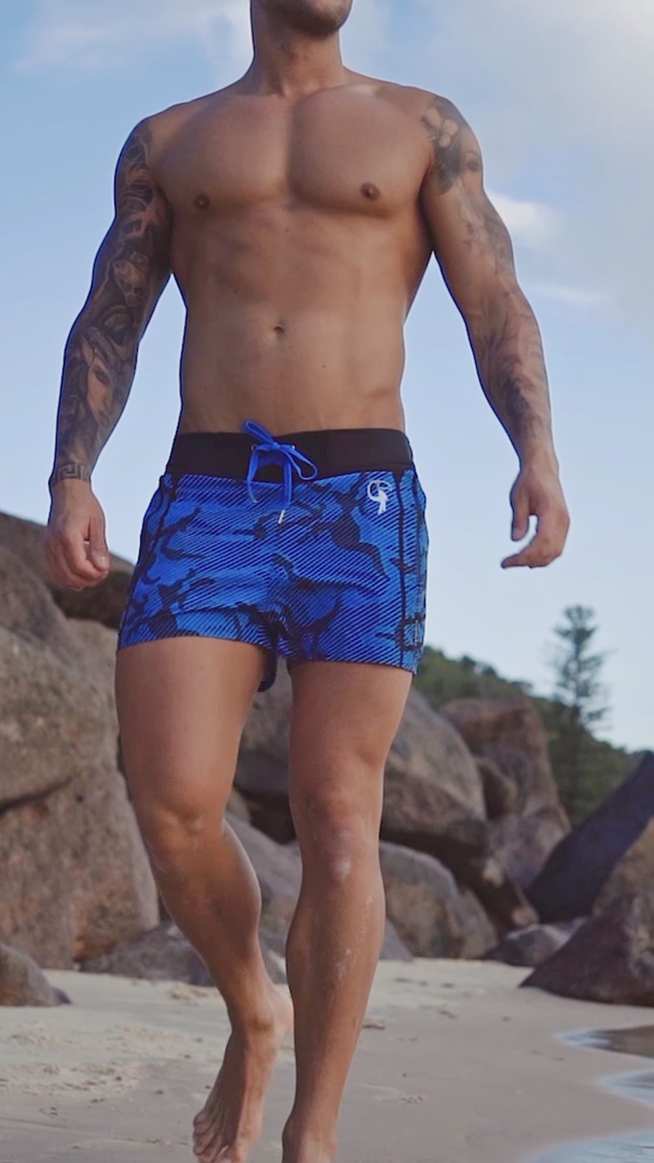 Man in striped camo blue swim shorts walking along a rocky beach.